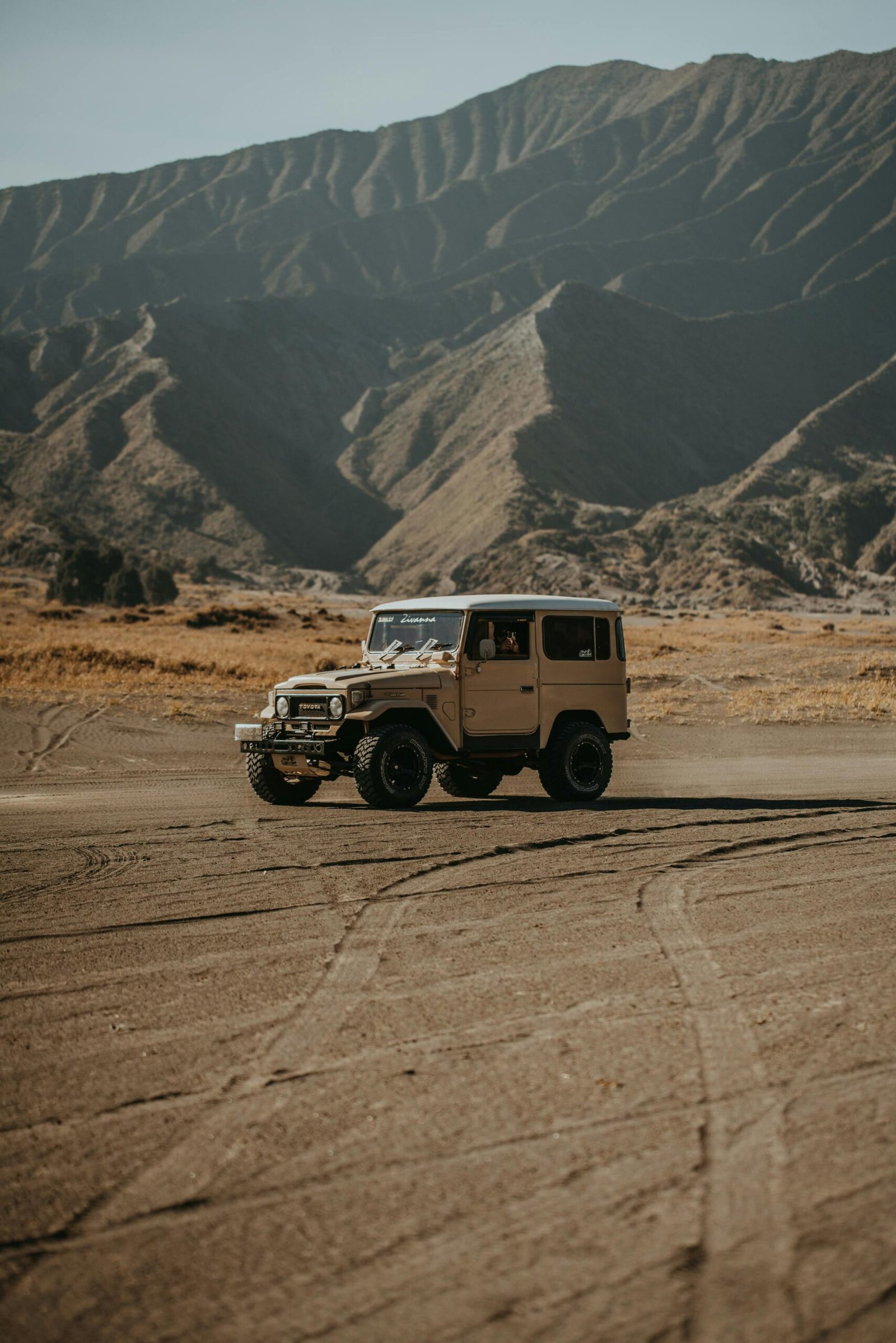 Vintage beige off-road Jeep driving across a dusty desert plain with rugged mountains in the background.

